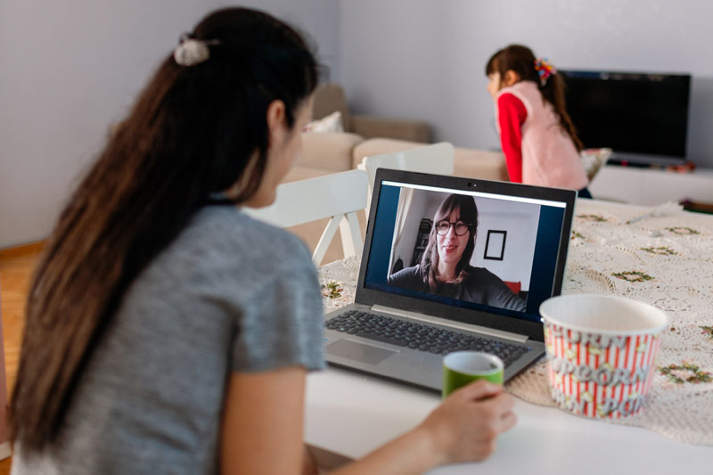 Photo of a girl holding a coffee mug and looking down at a video call with another person on her laptop
